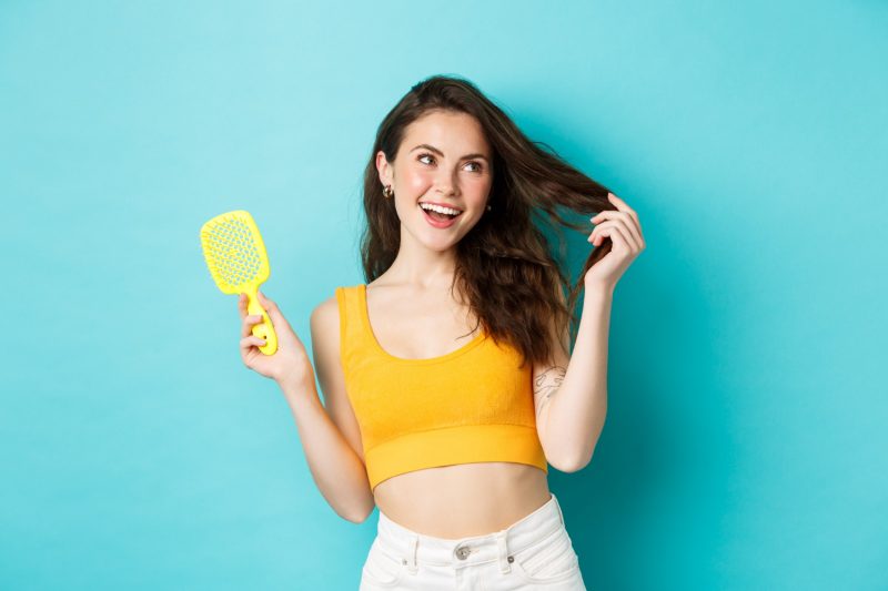 happy attractive woman showing her brush without hair strand touching healthy long hairstyle with pleased face smiling delighted with shampoo result blue background