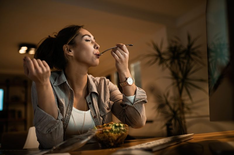 low angle view young woman eating salad with her eyes closed while using computer night home